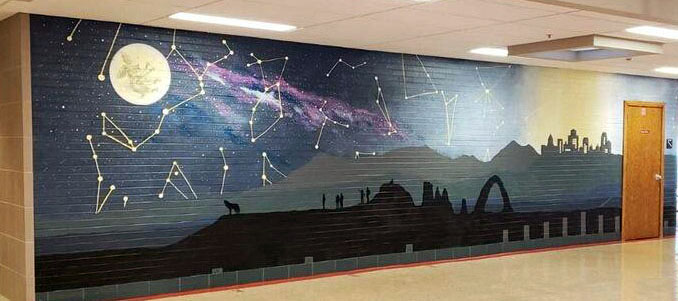 A full wall mural. From the left, the sky is dark and the stars and Milky Way are bright. There's a full moon with a figure our a rabbit in it. as the mural goes to the left, it gets closer to a city and the light pollution gets worse, obscuring the celestial bodies in the sky. There's a silhouette of a hill with a wolf and figures looking at the sky.