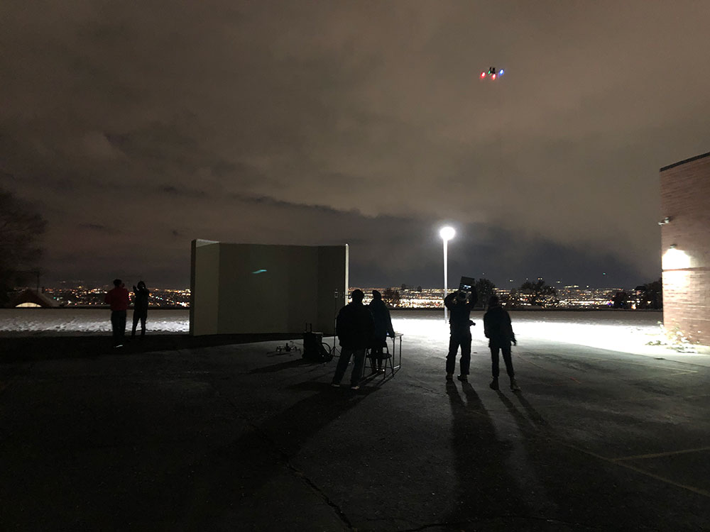 At night outside, six figures look up at an atonomous drone that's flying around a school taking brightness measurements. There's a very bright light, and beyond the school there are the bright lights on the city. The sky is cloudy.